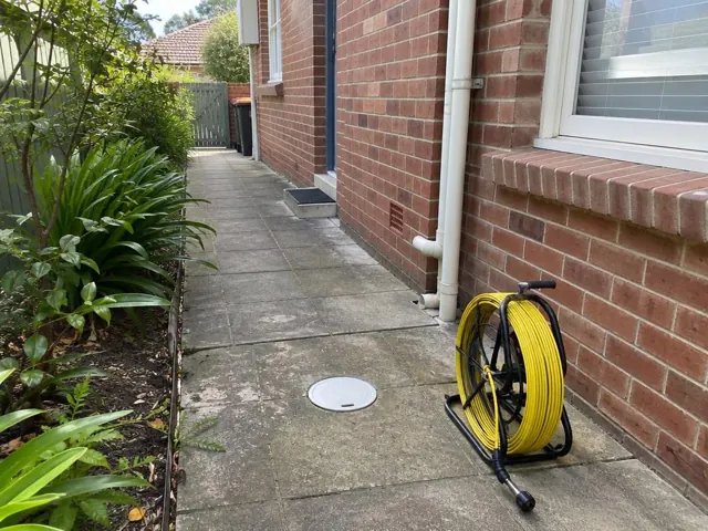 Tidy brick house side path with a capped drain inspection opening and a yellow camera reel beside it in Eastwood