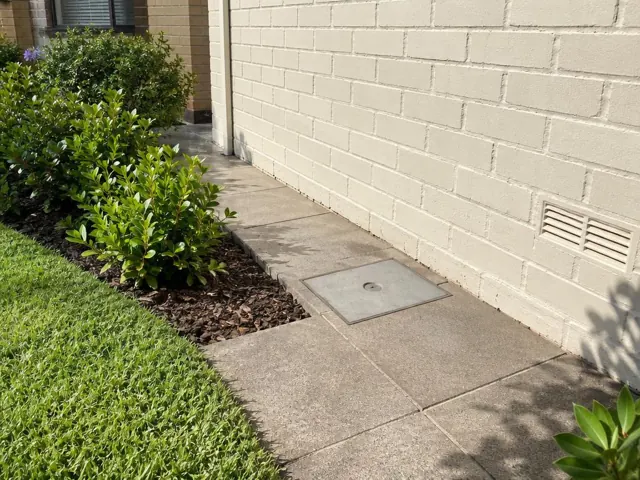 Flush square access cover in a paved side yard beside a brick house wall in Macquarie Park
